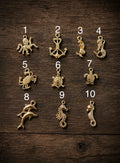 Ten small metallic jewelry charms arranged on a dark wood background, numbered 1 through 10. The charms include an octopus, anchor, mermaids, turtle, crab, dolphins, seahorses, and a sea turtle figure.