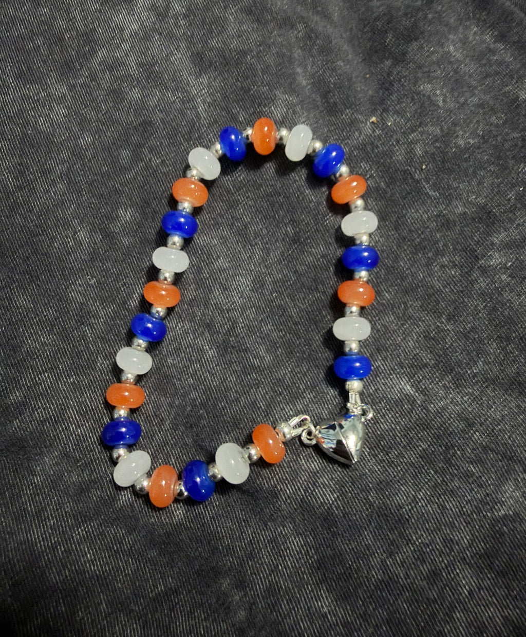 A beaded bracelet featuring alternating blue, orange, and white beads, separated by small silver spacer beads, with a silver heart magnetic closure at the clasp, displayed on a dark gray textured fabric background.