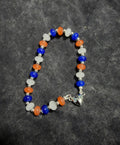 A beaded bracelet featuring alternating blue, orange, and white beads, separated by small silver spacer beads, with a silver heart magnetic closure at the clasp, displayed on a dark gray textured fabric background.