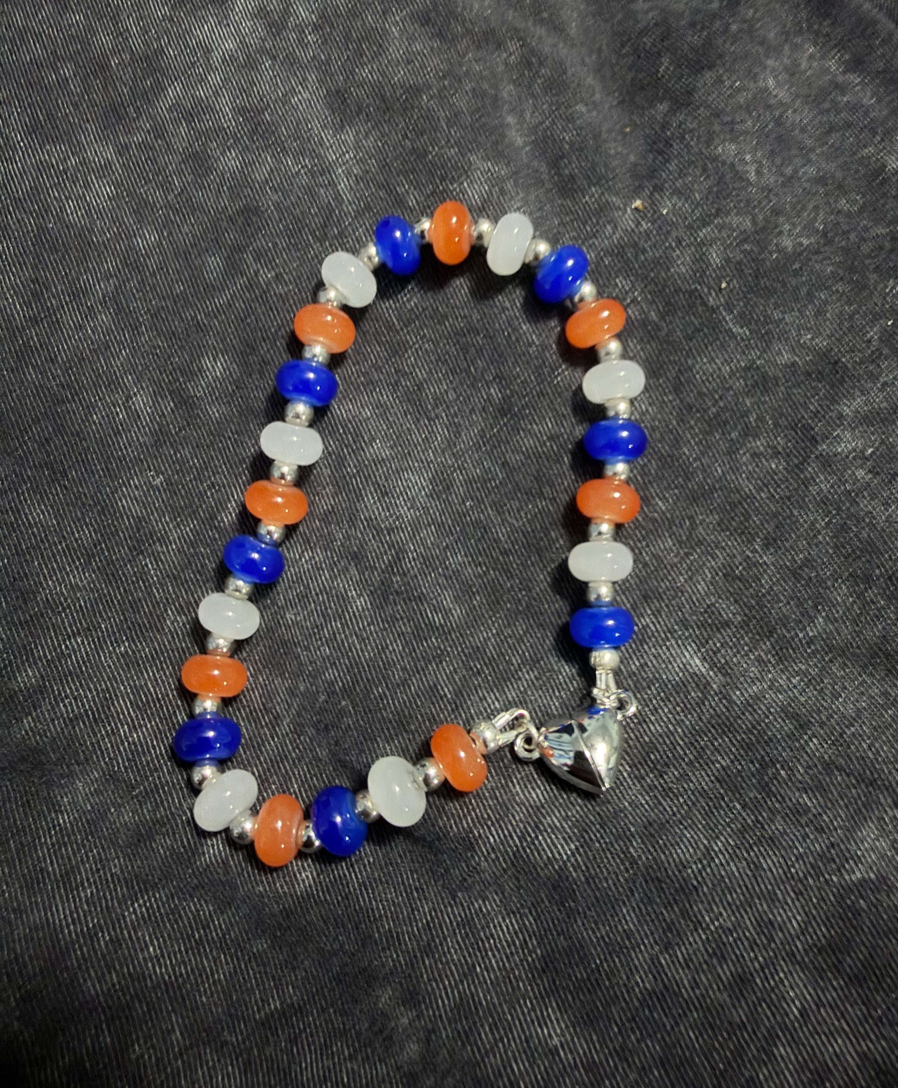 A beaded bracelet featuring alternating blue, orange, and white beads, separated by small silver spacer beads, with a silver heart magnetic closure at the clasp, displayed on a dark gray textured fabric background.