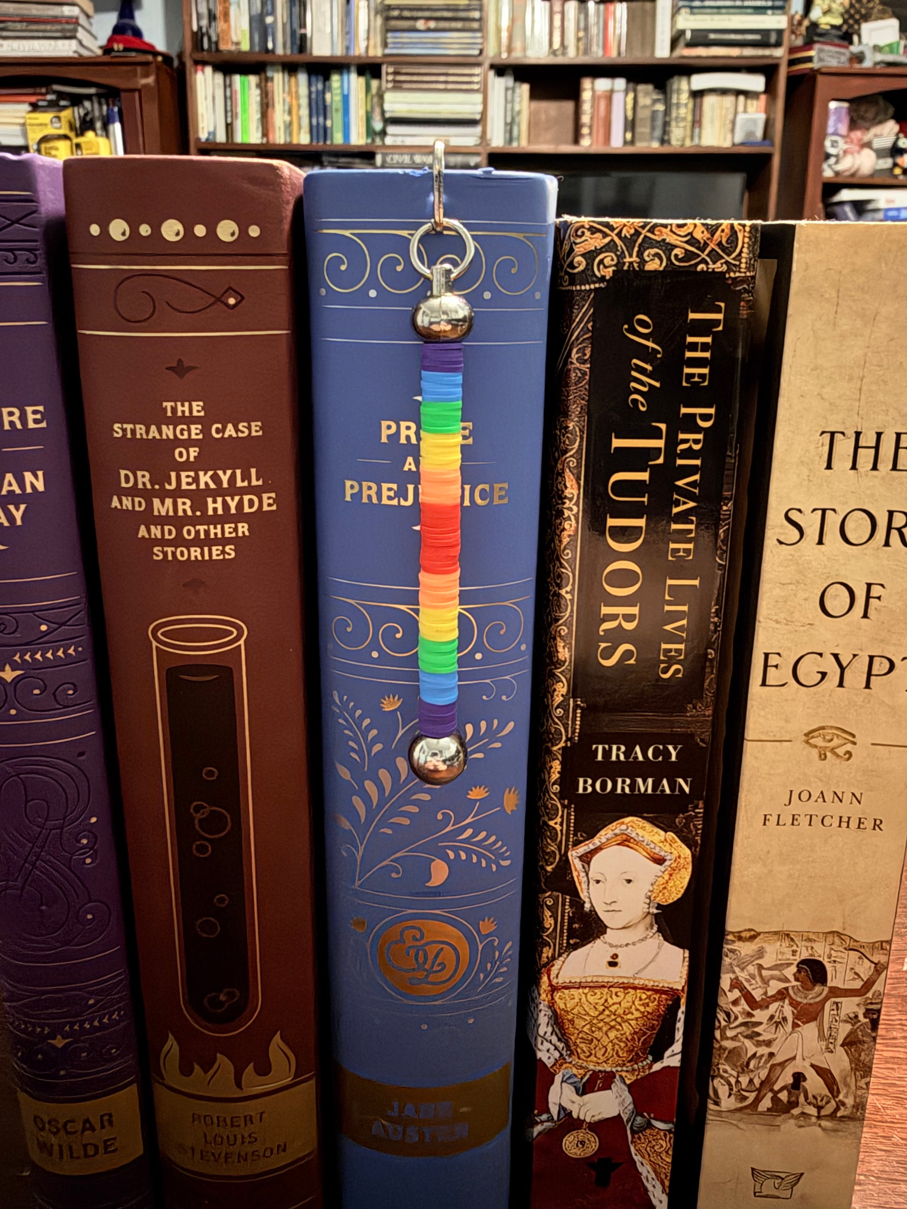 A rainbow-colored metal book bookmark with a small silver bell hangs from the spine of a blue book on a bookshelf, surrounded by other vintage books.
