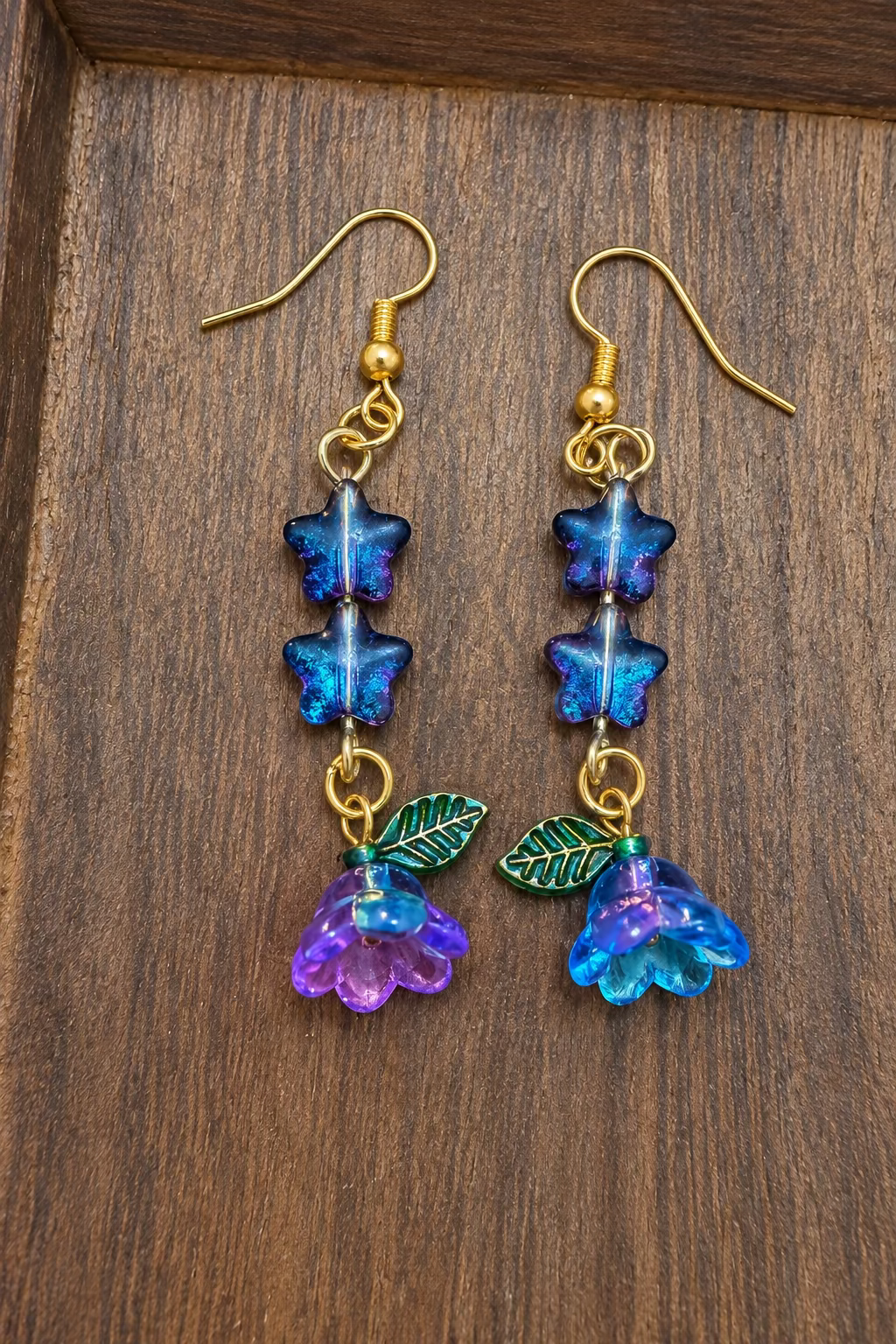 A pair of handmade dangle earrings displayed on a dark wooden surface. The earrings feature gold-tone fishhook wires, iridescent blue and purple star beads, a small green leaf charm, and a dangling flower charm that shifts between purple, pink, and blue. Note - the blue and purple in the stars is not normally that prominent - I had to use a flashlight and a camera flash to bring it out that boldly.