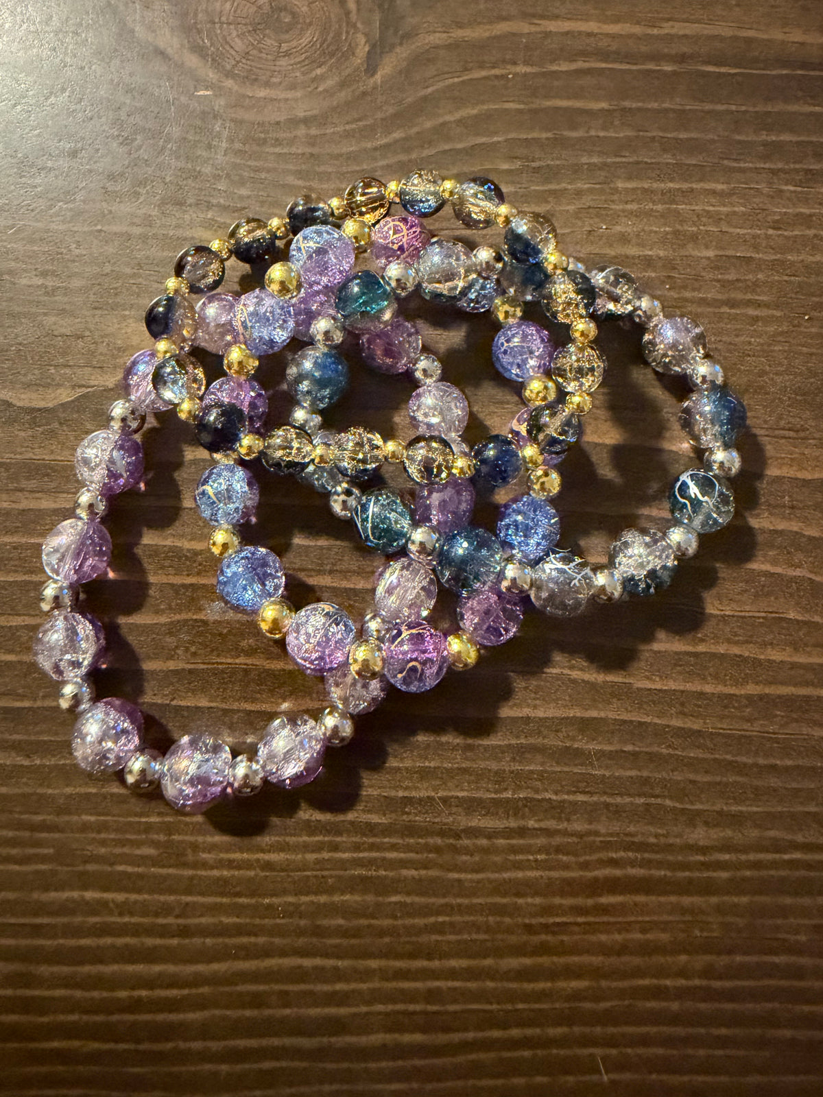 A set of beaded stretch bracelets with crackle glass beads in purple, blue, pink, and clear, separated by small metal spacer beads, displayed on a dark wood surface.
