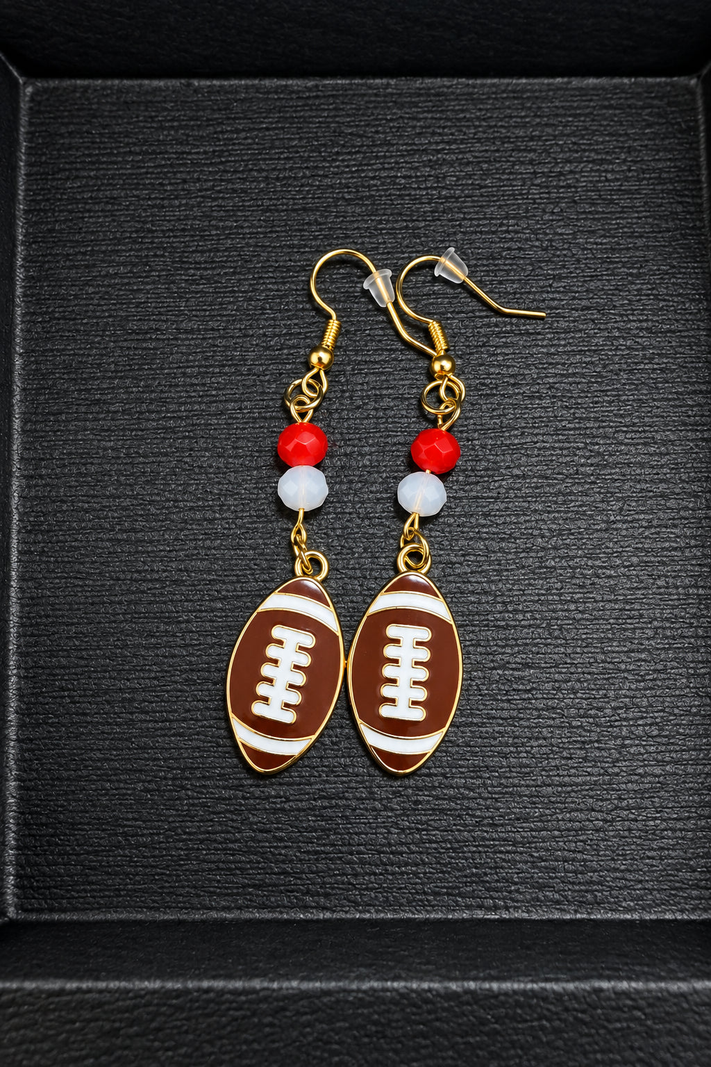 A pair of dangling earrings with football-shaped charms, red and white beads, and gold hooks, displayed on a dark wood surface.