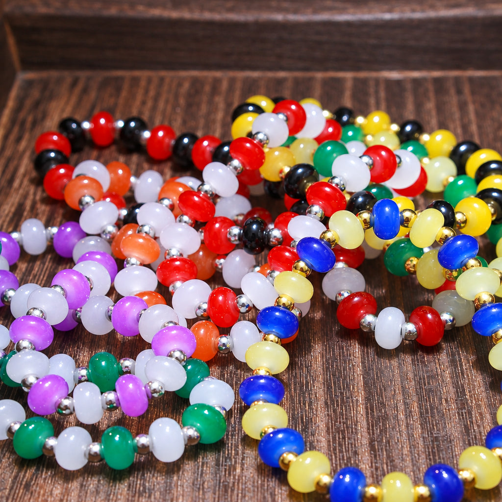 A close-up of multiple colorful beaded bracelets arranged on a dark wooden surface. The bracelets feature translucent round beads in red, black, white, purple, green, yellow, blue, and orange, separated by small metallic spacer beads.