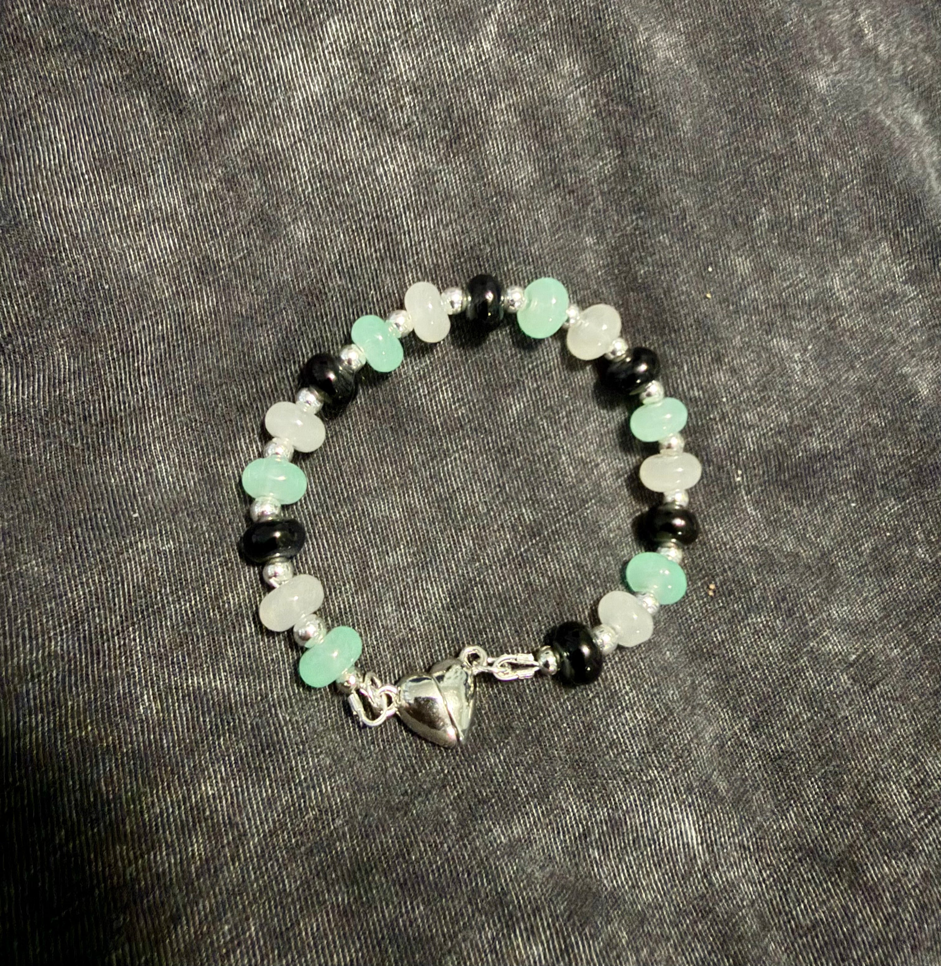 A beaded bracelet with alternating black, grey, and light green round beads, featuring a silver-toned heart-shaped clasp, displayed on a dark gray textured fabric background.