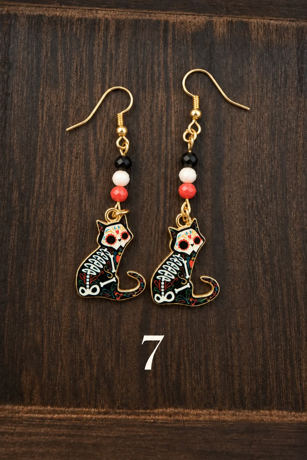 A pair of dangling earrings with gold-tone hooks and beads, featuring small black cat skeleton charms with white and red details, displayed on a dark wood background.