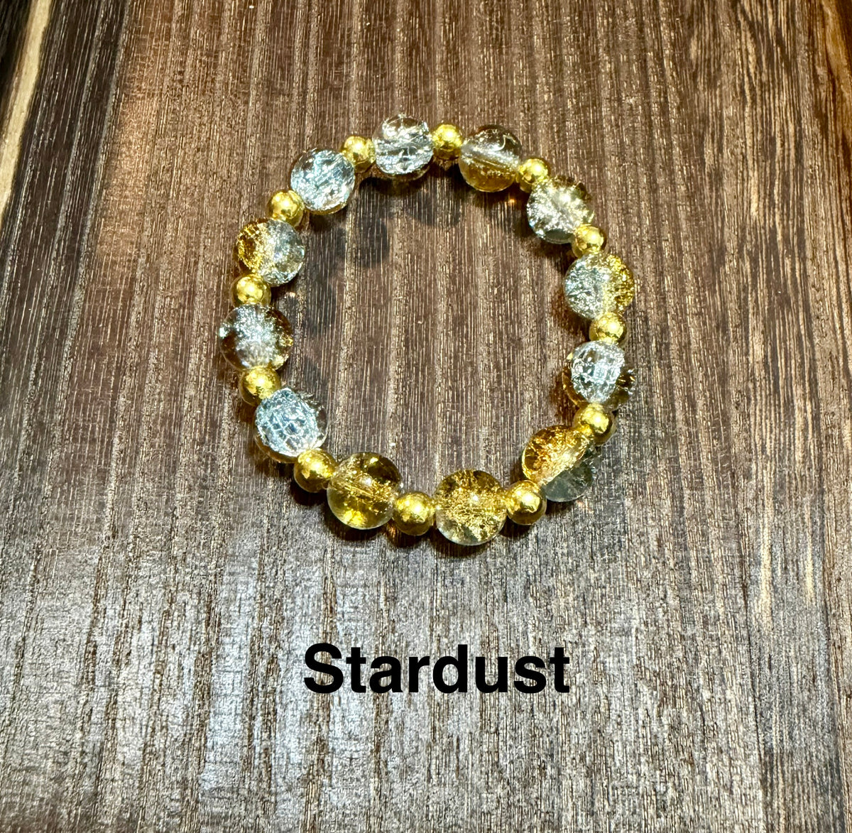 A beaded stretch bracelet featuring crackle glass beads showing gold and silver with small hints of sky blue and small gold spacer beads, displayed on a dark wood background with the text 'Stardust' underneath.