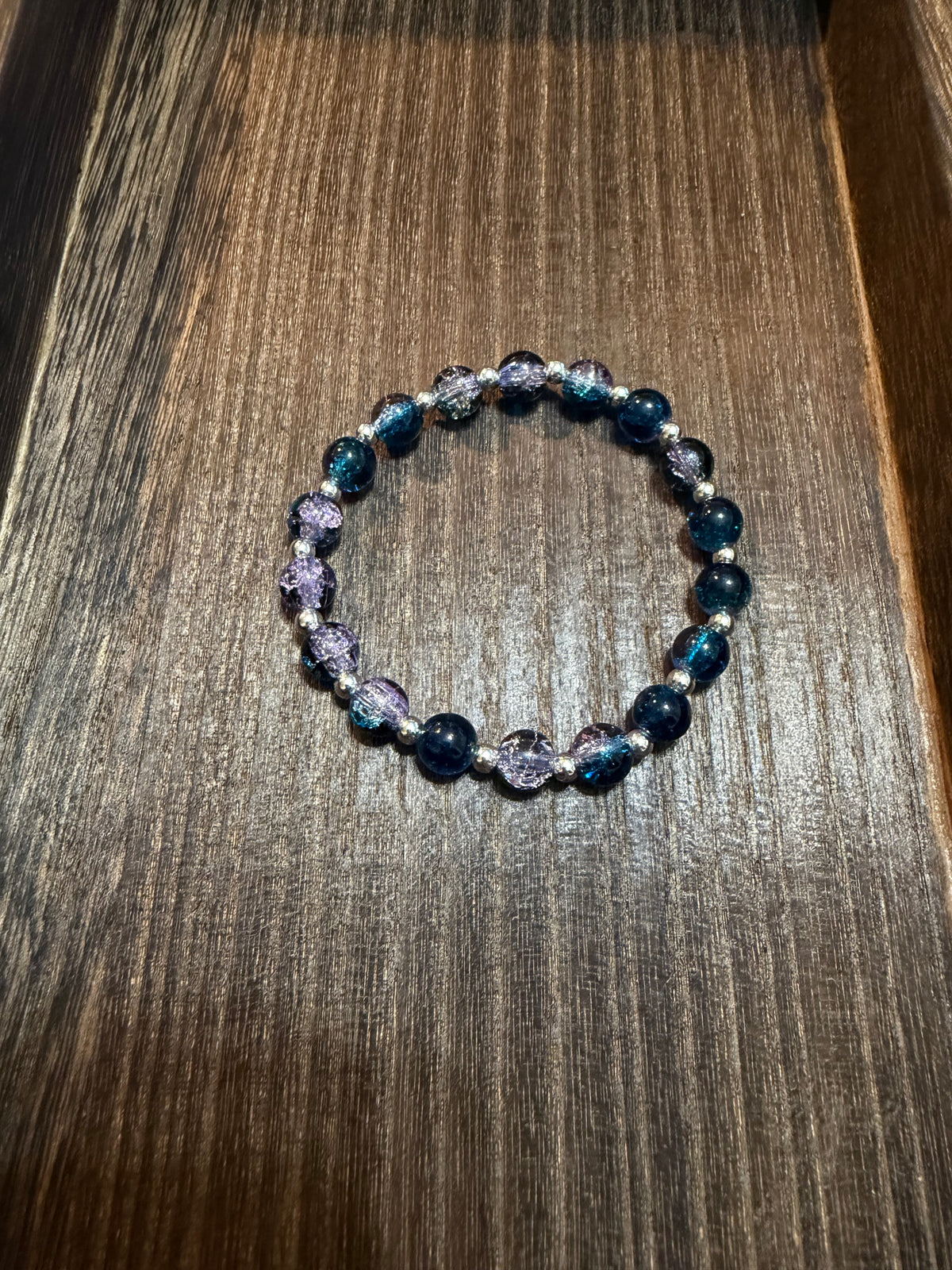 A beaded stretch bracelet featuring dark blue and lavender crackle glass beads with small silver spacer beads, displayed on a dark wood surface.