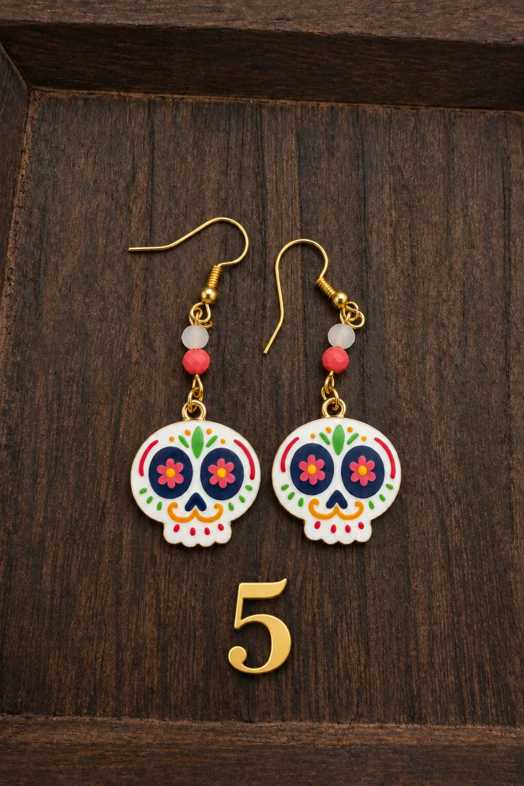 A pair of sugar skull drop earrings with gold hooks, red and white beads, and colorful painted skulls, displayed on a dark wood background.