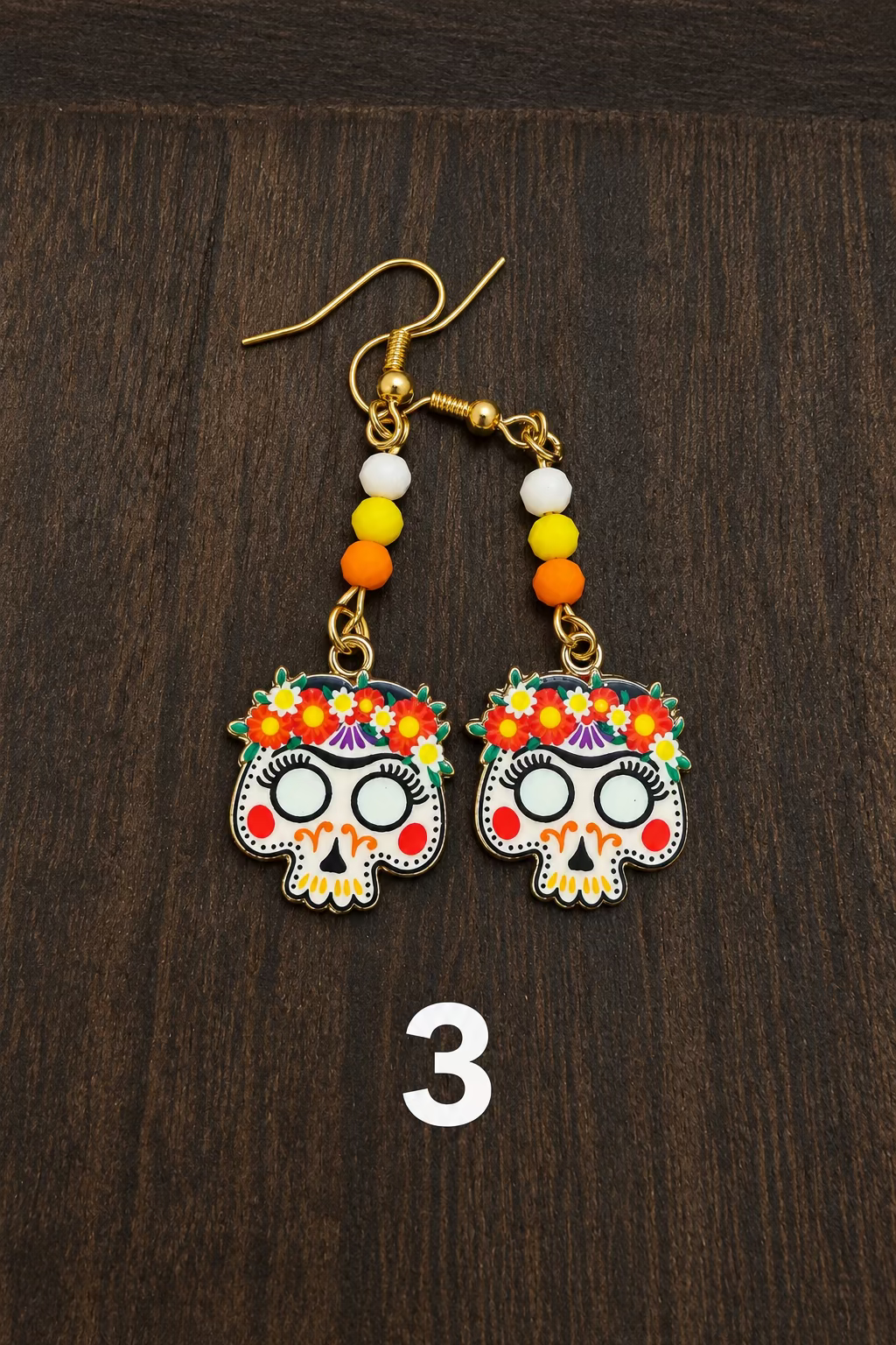 A pair of dangling earrings with gold fishhook wires, small white, yellow, and orange beads, and colorful floral sugar skull charms, displayed on a dark wood surface.