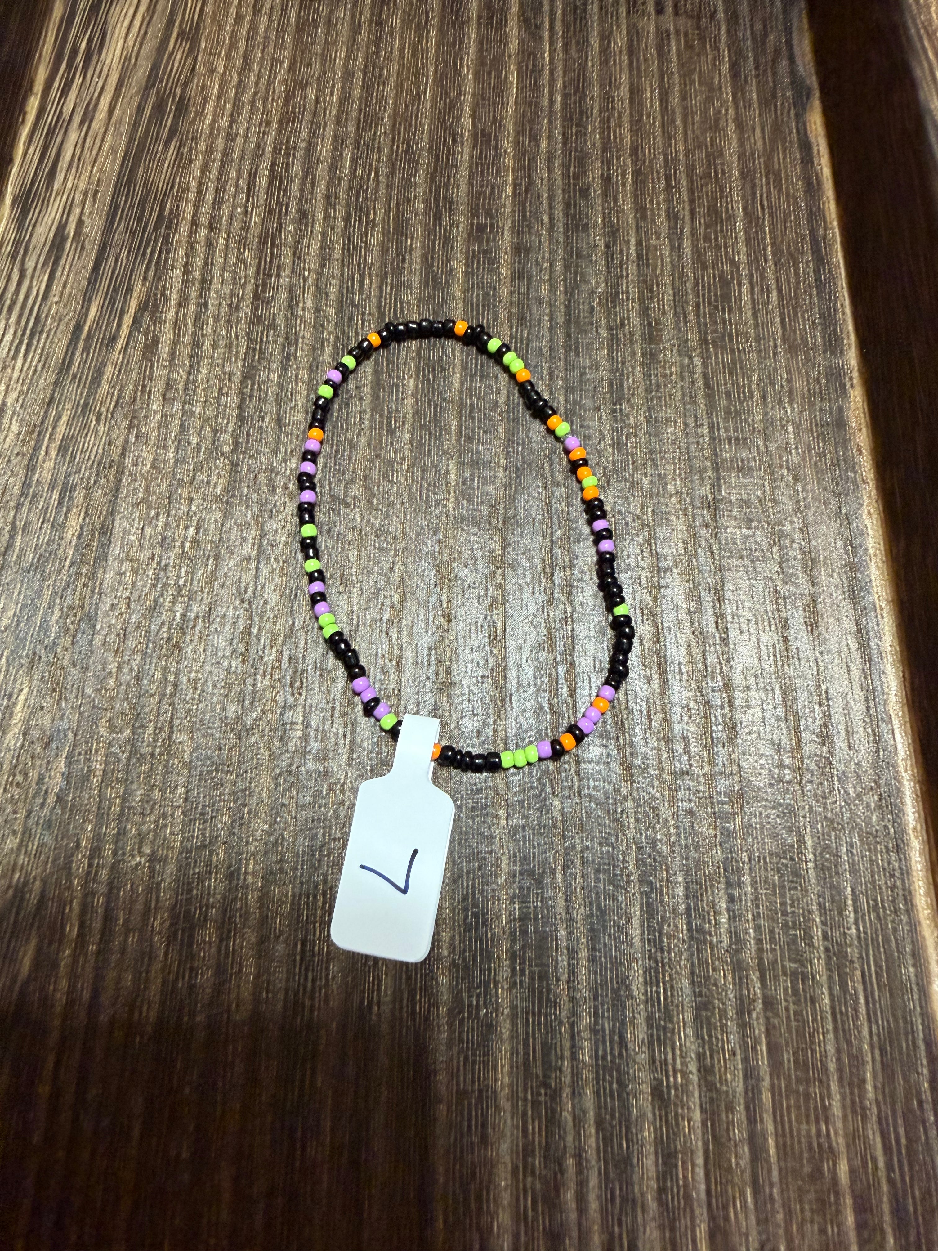 A beaded bracelet displayed on a dark wood surface. The bracelet features small multicolored beads, primarily black, with orange, green, and purple, with a white tag marked with a black checkmark.