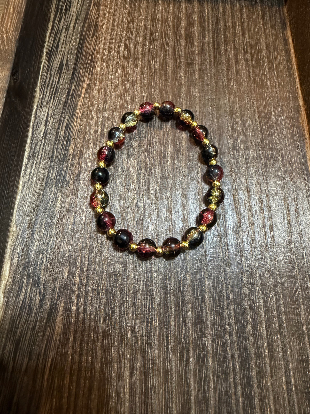 A beaded stretch bracelet featuring crackle glass beads in dark green, dark pink, and gold, arranged in a circle on a dark wood surface.