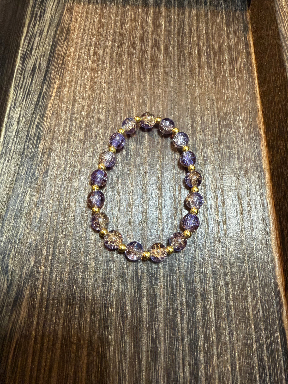 A beaded stretch bracelet made of purple, gold, and silver crackle glass beads separated by small gold spacer beads, displayed on a dark wood surface.