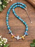 A beaded necklace with turquoise-blue Aquamarine stone beads, small clear faceted beads, and colorful fish-shaped beads in blue, orange, yellow, and silver, displayed on a rustic wooden surface with small white flowers.