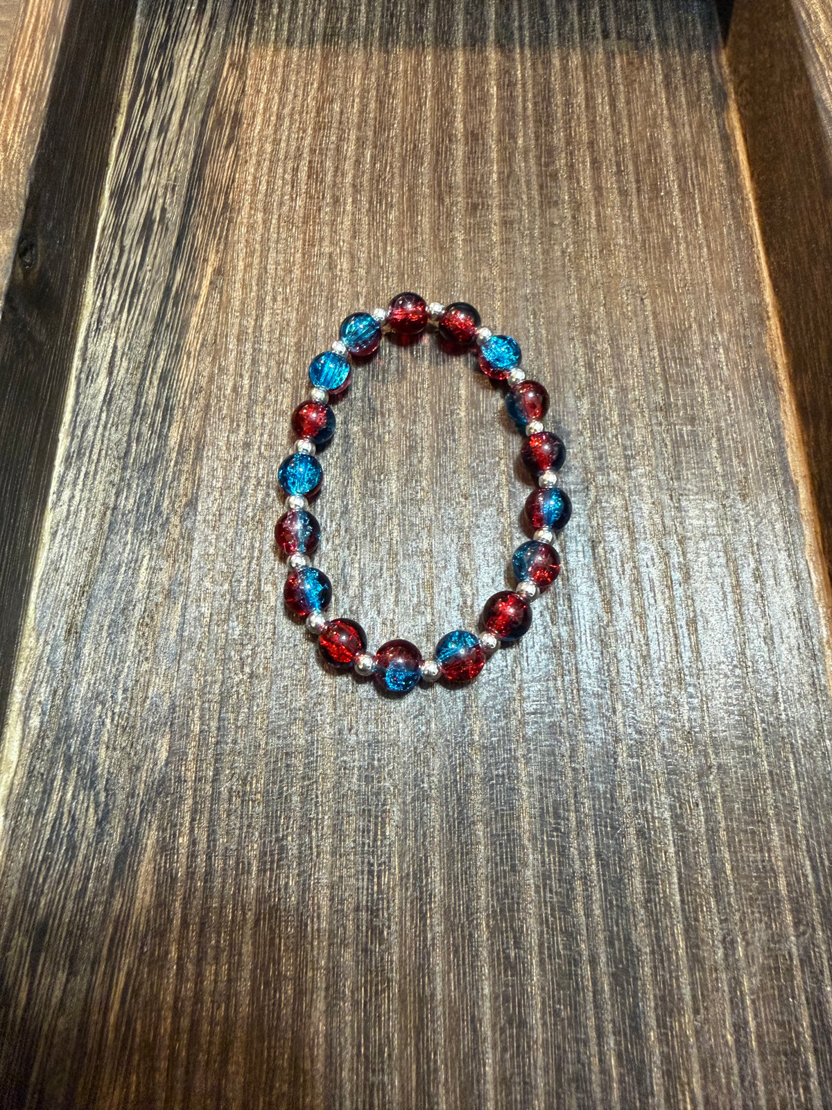 A beaded stretch bracelet with red and blue crackle glass beads and silver spacer beads, displayed on a dark wood surface.