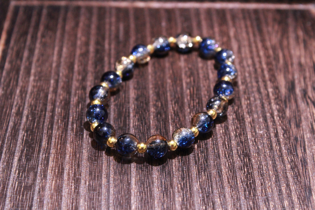 A beaded stretch bracelet featuring dark blue, clear, and gold crackle glass beads separated by small gold spacer beads, displayed on a dark wood surface.