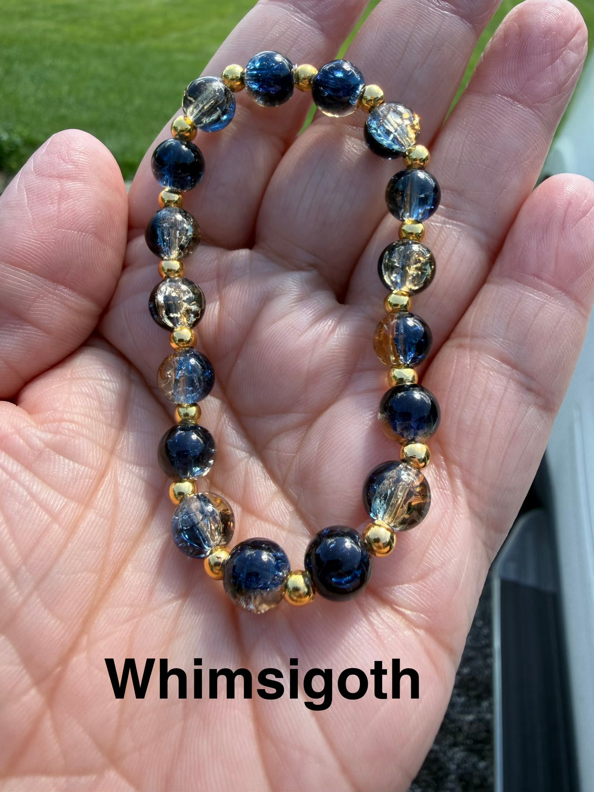 A beaded stretch bracelet held in a hand, featuring crackle glass dark blue, clear, and gold beads and small gold spacer beads. The text 'Whimsigoth' is overlaid at the bottom.  It is inspired by the whimsigoth style of the late 90s.