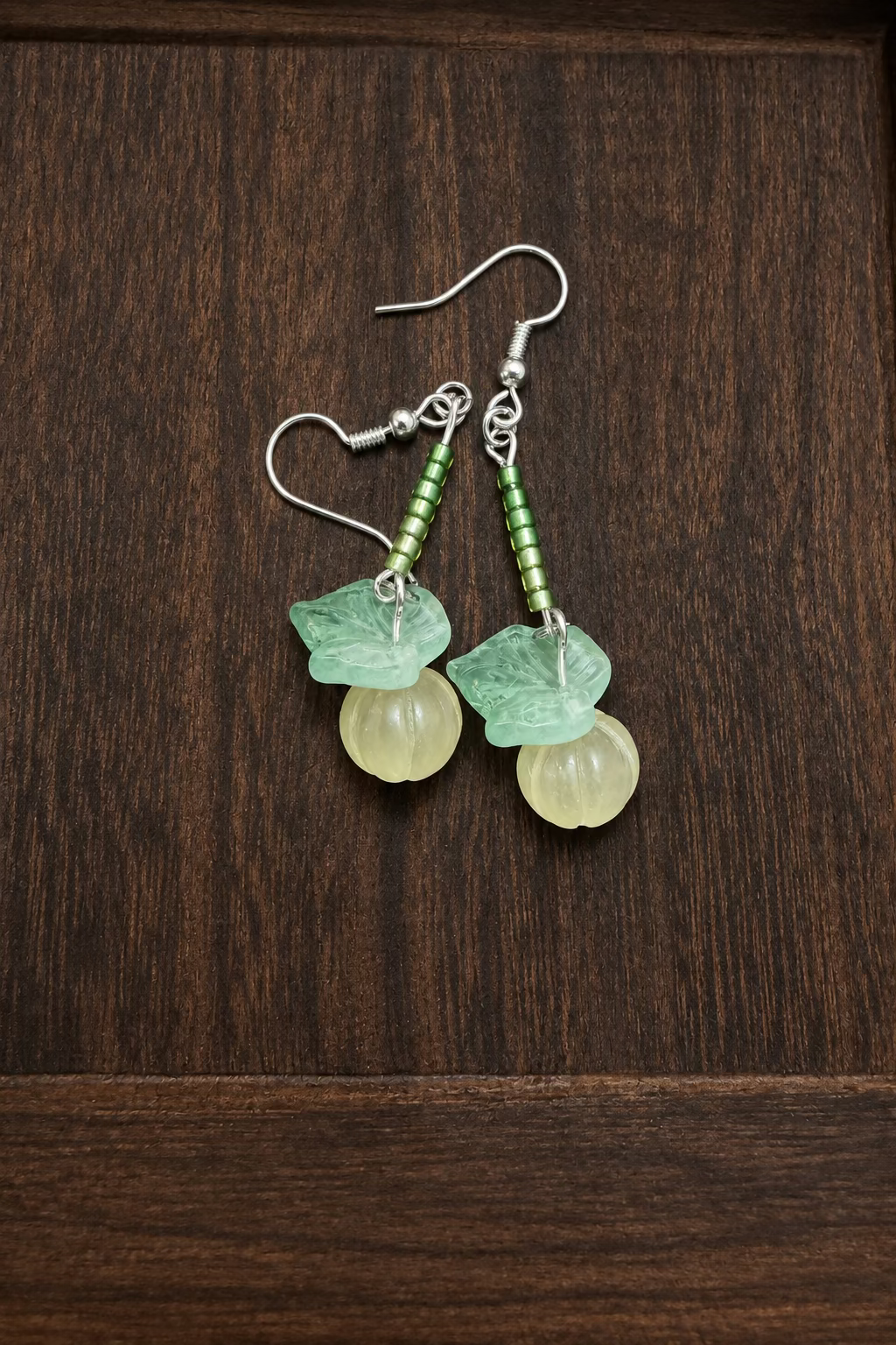 A pair of handmade dangle earrings featuring light green glass leaves and small green seed beads above white carved stone pumpkins displayed on a dark wood surface. 
