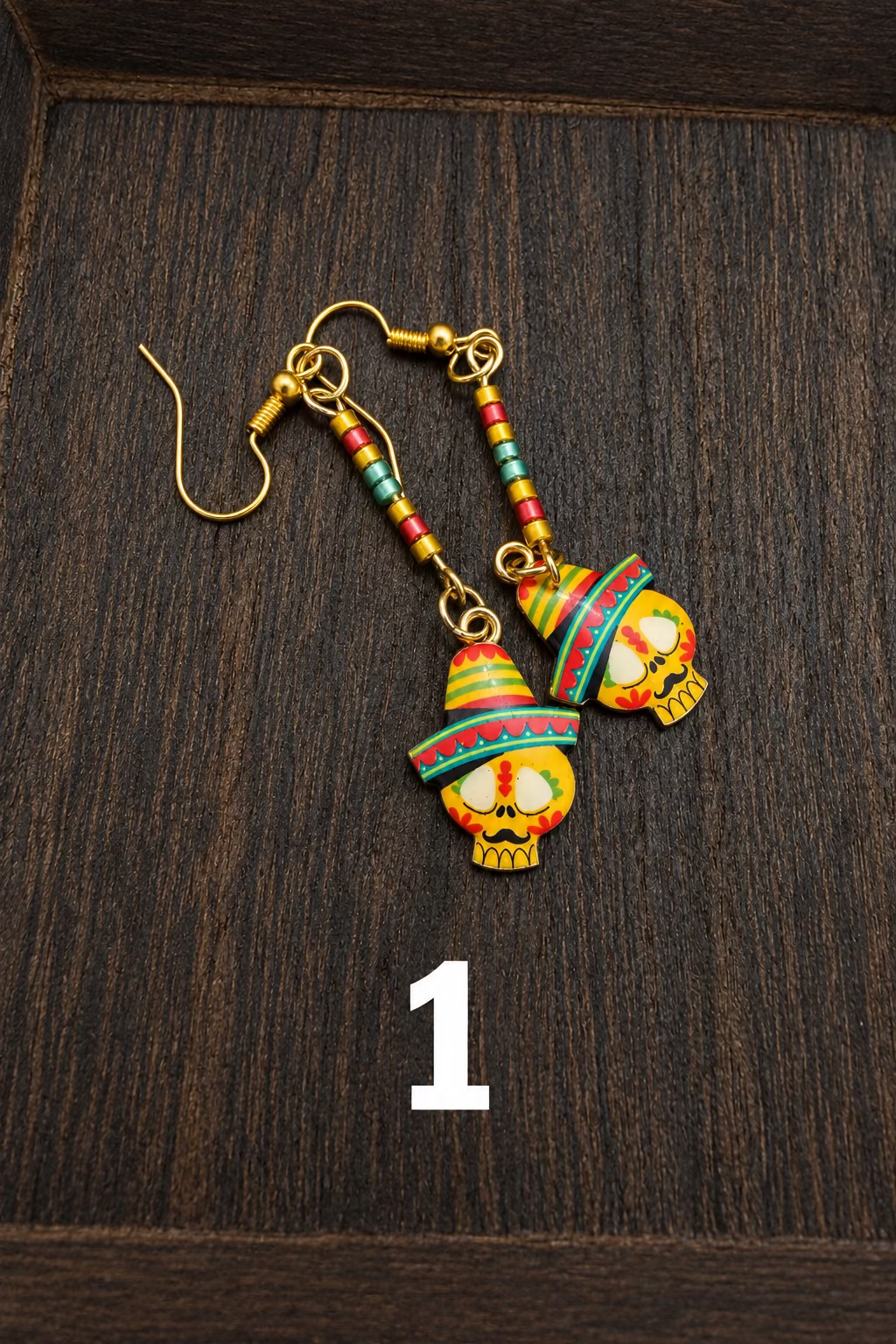A pair of dangling earrings with gold-tone hooks and glass beads, featuring colorful skull charms wearing red, green, and yellow striped sombreros, displayed on a dark wooden surface.